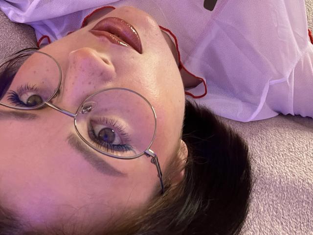 MedeaAllure's face, lying down, with glasses, purple eyelids, and tongue slightly out, wearing a white shirt with red trim, on a textured gray surface.