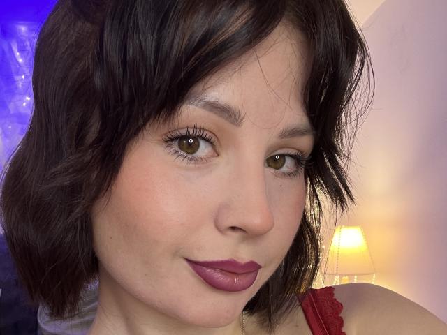 MedeaAllure has short brown hair, green eyes, and pink lipstick. She's smiling slightly with her upper body in a red top. Her tits are not visible. Lamp in background.