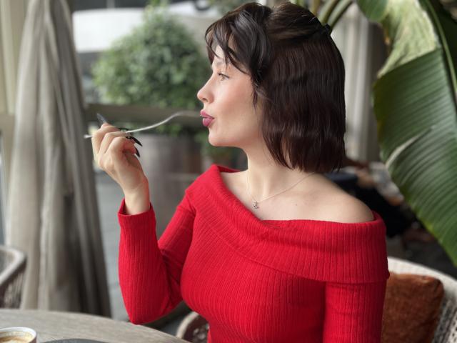 MedeaAllure with short brown hair, red off-shoulder sweater, eating with a fork, her tits slightly visible, looking to the side, sitting at an outdoor table with a plant.