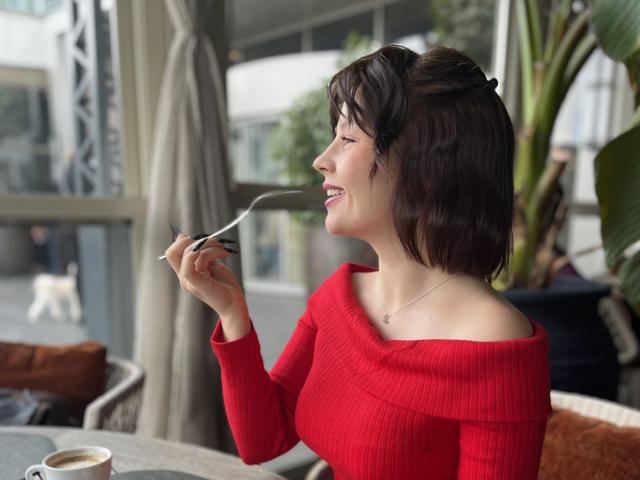 MedeaAllure with short brown hair, red off-shoulder sweater, holding spoon to mouth, smiling, sitting at table with coffee, outdoor setting, green plant in background, sunlight.