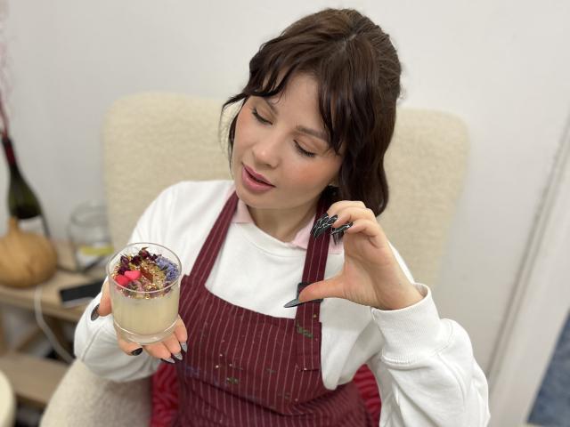 MedeaAllure with brown hair, wearing a white shirt and red pinstriped apron, holding a glass of dessert, showing long black nails, looking down, in a cozy room.