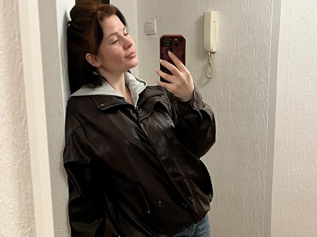 MedeaAllure with brown hair, wearing a black leather jacket and blue jeans, taking a mirror selfie with a phone, eyes closed, slightly pouty expression, white wall and phone behind.