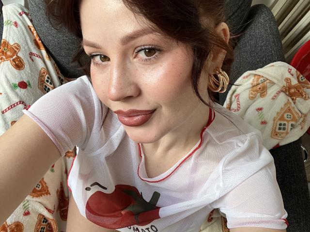 MedeaAllure with fair skin, brown eyes, and red lipstick, wearing a white shirt with a red heart, gold hoop earrings, and a Christmas-patterned blanket, smiling at the camera.
