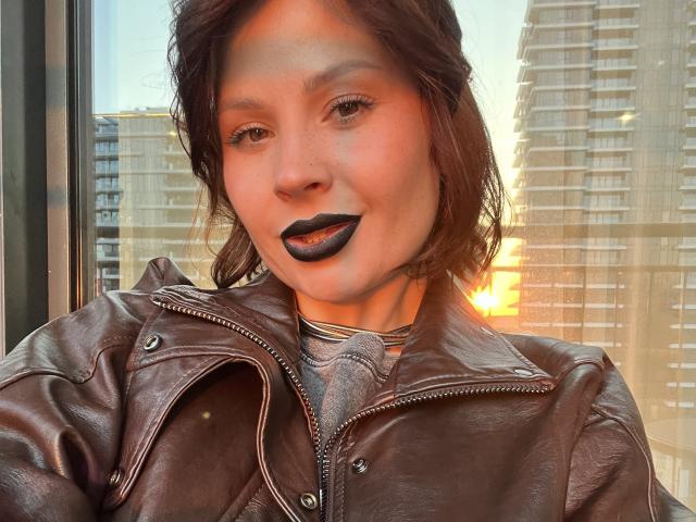 MedeaAllure with dark lipstick, brown leather jacket, and wavy hair, standing by a window at sunset, city buildings in the background, warm light highlighting her face.