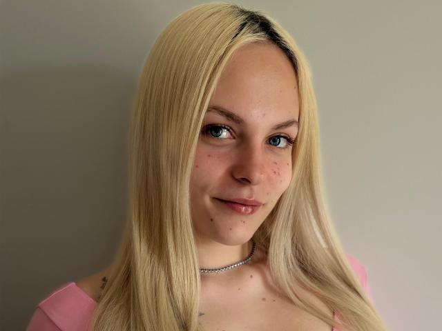 ZayaIzBack has blonde hair, blue eyes, and is smiling with a slight nose and lip piercing. She's wearing a pink top and a silver necklace.