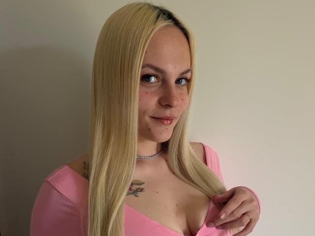 ZayaIzBack tem cabelos loiros longos, olhos azuis e um pequeno sorriso. Ela está vestindo uma blusa rosa, que mostra seus seios, com uma tatuagem acima do seio esquerdo. Colar de choker..
