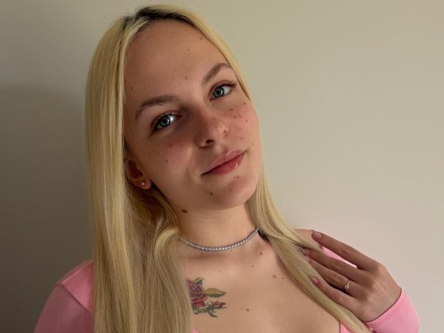 ZayaIzBack hat blondes Haar, blaue Augen und ein kleines Rosentattoo auf ihrer Brust. Sie trägt ein pinkes Oberteil, eine Halskette und einen Ring an ihrer rechten Hand..