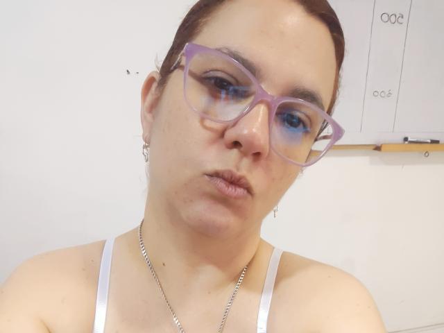 AmeliaWilson has puffy purple glasses, brown hair in a bun, wearing a white tank top, and a silver chain, with a neutral expression and slightly pursed lips.