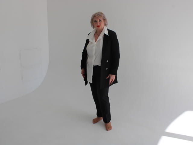 RenataDaster stands barefoot, wearing a black jacket, white shirt, black pants, and brown shoes, with short blonde hair and a red lipstick smile, in a white-walled studio.