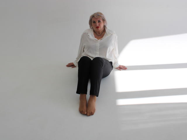 RenataDaster with pale blonde hair, red lipstick, white button-up shirt, black pants, bare feet, sitting on white floor, arms slightly behind her, shadow on right.