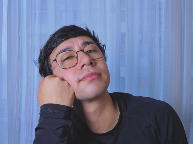 OliverHot69 with black hair, glasses, and a slight smile, resting his head on his hand, wearing a black shirt, looking relaxed and slightly cute.
