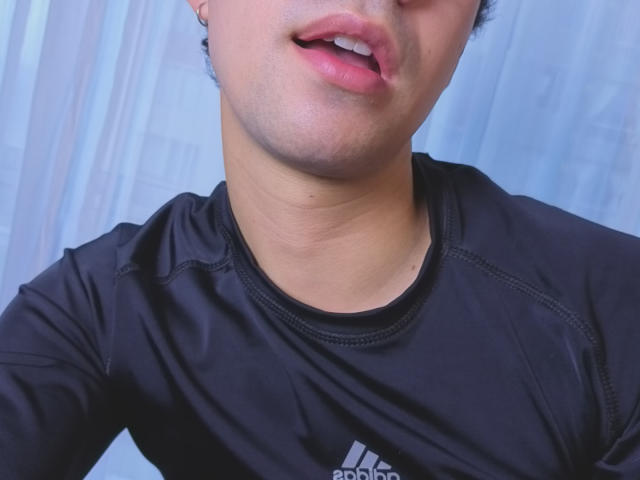 OliverHot69's face with slightly parted pink lips, wearing a black Adidas shirt, showing a small part of his chin and neck, with a light blue background.
