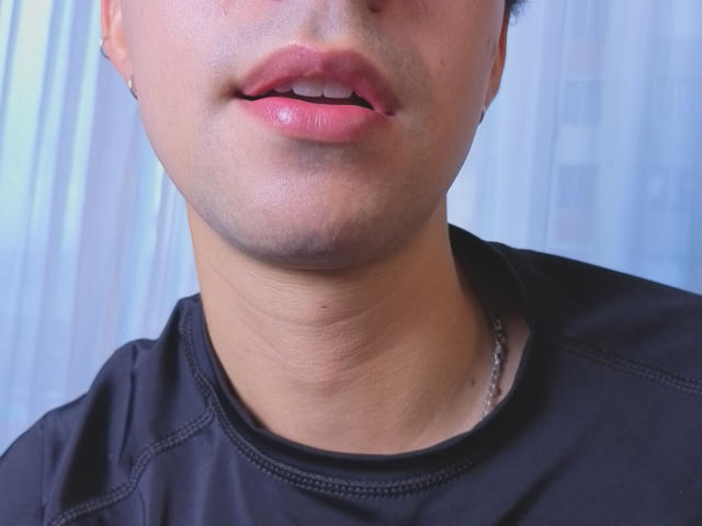 OliverHot69's lower face, showing slightly parted pinkish lips with a small hole in the upper lip, wearing a black shirt and a silver chain.