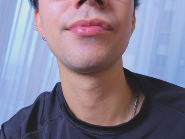 OliverHot69's face, showing a slight pout with glossy pink lips, smooth light brown skin, and a small hoop earring in his left ear, wearing a black shirt.