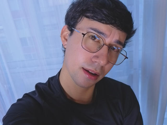 OliverHot69 with glasses, short black hair, black shirt, light skin, slightly open mouth, looking at camera, blue-toned background.