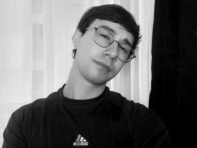 Black-and-white photo of OliverHot69 with short dark hair, glasses, slight smirk, black Adidas shirt, and ear piercings. OliverHot69's head is tilted to the side.