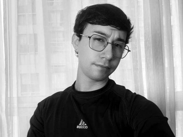 OliverHot69 has short black hair, glasses, and a slight smirk. He's wearing a black Adidas shirt, and his ear has a small hoop earring. Black-and-white photo.