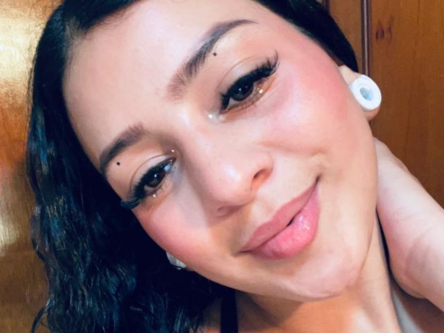 LonxEva with medium brown skin, black wavy hair, thick eyebrows, small beauty mark under her right eye, and white earbuds, smiling with pink lips, has a small drop on her cheek.