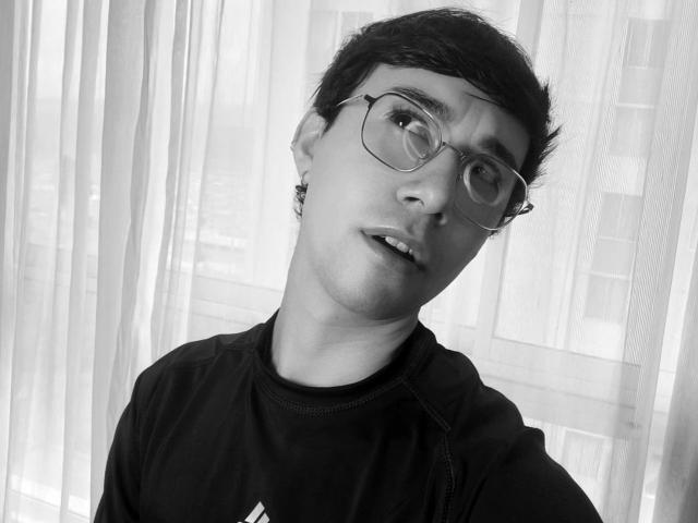Black-and-white photo of OliverHot69 with short dark hair, glasses, slightly parted lips, and a black t-shirt. OliverHot69's expression is curious and he's looking to the right.