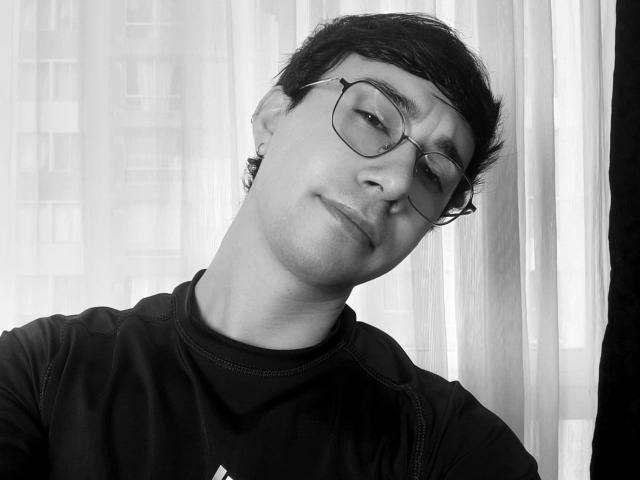 OliverHot69 with short dark hair, glasses, and a slight smirk, wearing a black shirt, has a small earring in his left ear. Black and white photo.