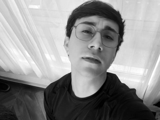 Black-and-white selfie of OliverHot69 with short dark hair, glasses, and a slightly confused expression. Wearing a dark shirt, small earring visible on left ear.