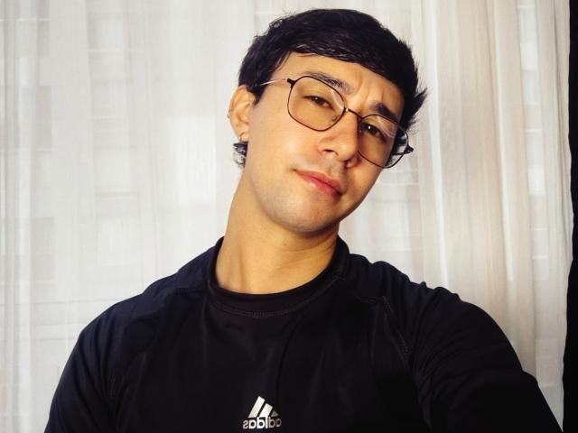 OliverHot69 has short black hair, glasses, and a slight smile, wearing a black Adidas shirt with the three stripes logo, in a selfie.