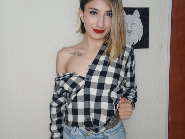 Kaiix with shoulder tattoos, wearing an off-shoulder black-and-white checkered shirt, blue jeans, red lipstick, smiling, blonde hair, one hand on shirt, standing in front of a wall.