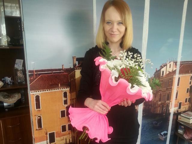Alisa, mit blonden Haaren, schwarzem Hemd, hält einen pinken Blumenstrauß mit weißen Blumen und Babys Atem, lächelnd. Ein pinkes Band ist aufgerollt. Sie steht vor einer Wandmalerei..