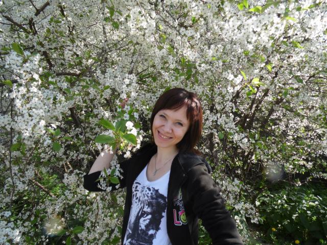 AlisaRogs with short brown hair, smiling, wearing a black jacket and white graphic tee, standing in front of a blooming white-flowered bush, holding a leaf.