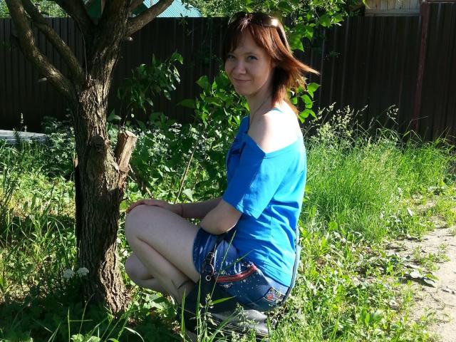 AlisaRogs seduta a gambe divaricate vicino a un albero, indossa una camicia blu con una spalla scoperta e pantaloncini di jeans, ha i capelli rossi, sta sorridendo, sullo sfondo si vedono l'erba verde e una recinzione marrone..