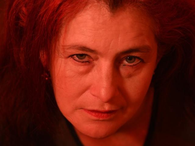 HelenaHank has red, messy hair, a slightly wrinkled face, green eyes, and a subtle, serious expression under red lighting. Her small nose and lips are clearly visible.