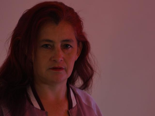HelenaHank has red hair, light skin, and small earrings, wearing a pinkish shirt with white trim, looking slightly annoyed in soft red lighting.