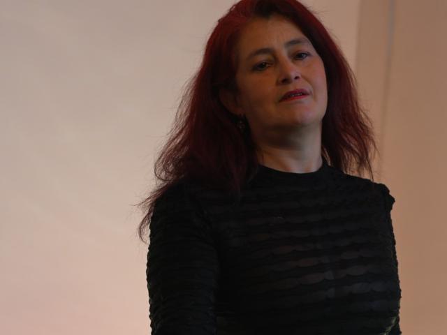 HelenaHank with red hair, wearing a black, sheer, textured top, smiling slightly, looking up, has small breasts, is standing, and has a light skin tone.