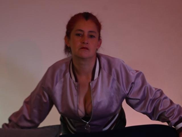 HelenaHank with red hair, wearing a shiny purple satin blouse, slightly open to show her tits, leaning forward with a serious expression, in dim, pinkish light.