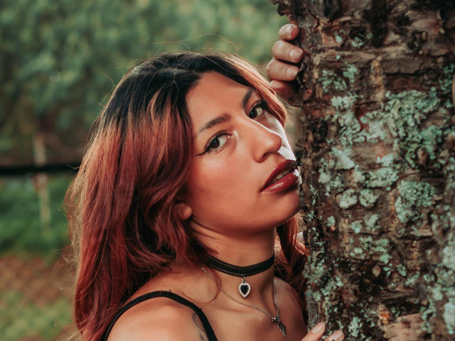 Alison, com cabelo ombré de vermelho para marrom, um colar preto e batom escuro, apoiada em uma árvore com textura, olhando para cima com uma expressão sedutora. Sua mão está agarrada à árvore..