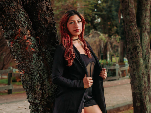 AlisonCold with red hair, black jacket, and black dress, standing against a tree, holding jacket open, showing black panties, choker necklace, and a serious expression.