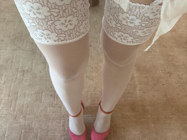 Tisia's white lace-topped thigh-highs, sheer white stockings, and pink heels with red straps, standing on a beige carpet, viewed from above. Her legs are visible, but not her ass.