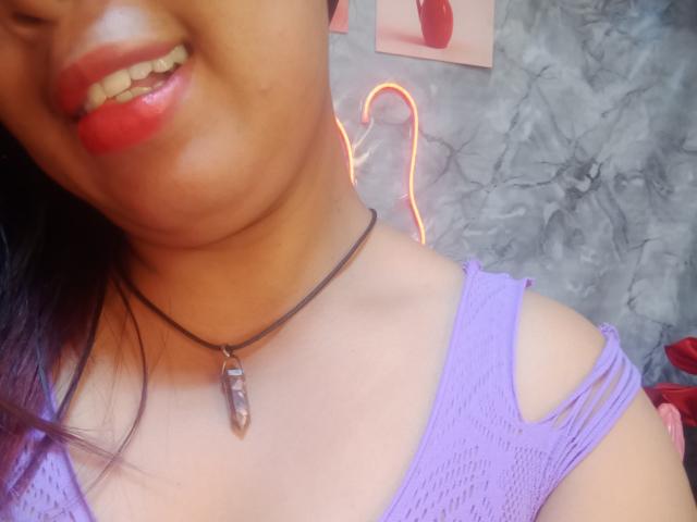MariahSquirting69 con lápiz labial rojo, una blusa de malla púrpura y un collar negro con un colgante de cristal, mostrando su parte superior del pecho y sus dientes, cabello púrpura y una pared de mármol gris..