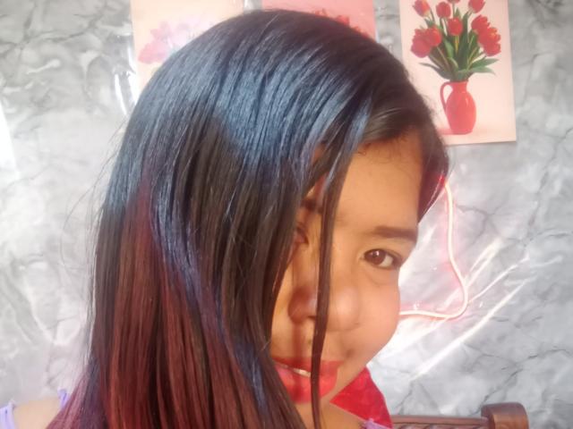 MariahSquirting69 a les cheveux noirs lisses avec des mèches rouges, des yeux marrons, et un collier rouge. Elle sourit légèrement et son nez est visible. L'arrière-plan comprend des peintures florales..