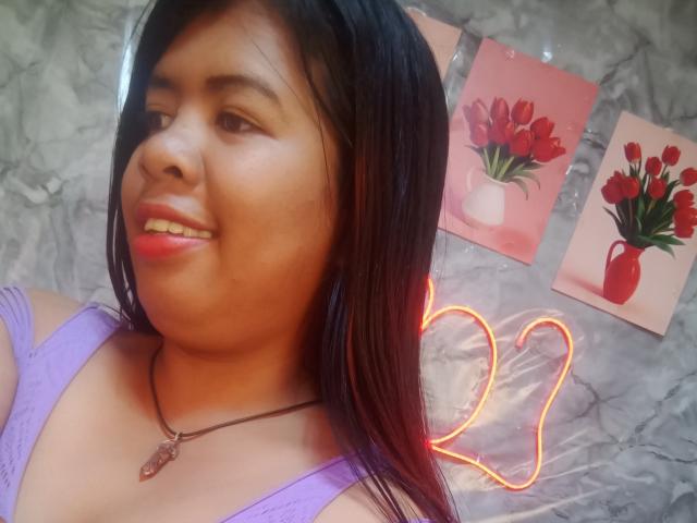 MariahSquirting69 with dark hair, light brown skin, purple top, necklace, smiling, pink lipstick, red eyes, two pink and red tulip paintings, neon heart on marble wall.