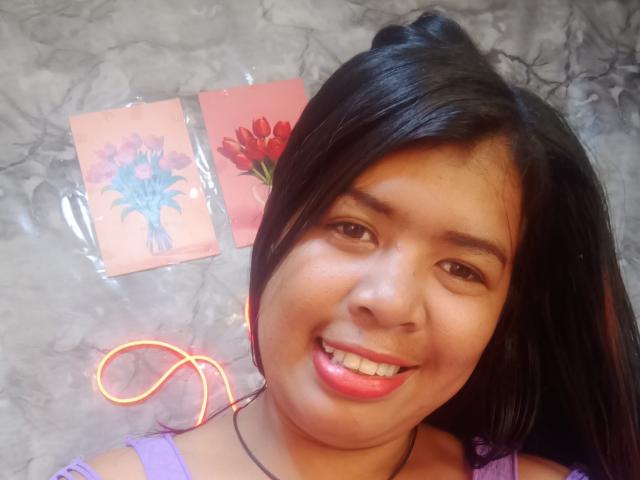 MariahSquirting69, uma mulher asiática atraente com cabelo preto, sorrindo com batom rosa, vestindo uma blusa roxa, e dois cartões coloridos em formato de flor dentro de sacos plásticos ao fundo..