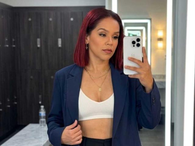 SharonBellarie with red hair, wearing a navy blazer, white bra, and gold necklaces, taking a mirror selfie, showing her tits, holding a white iPhone, water bottle nearby.