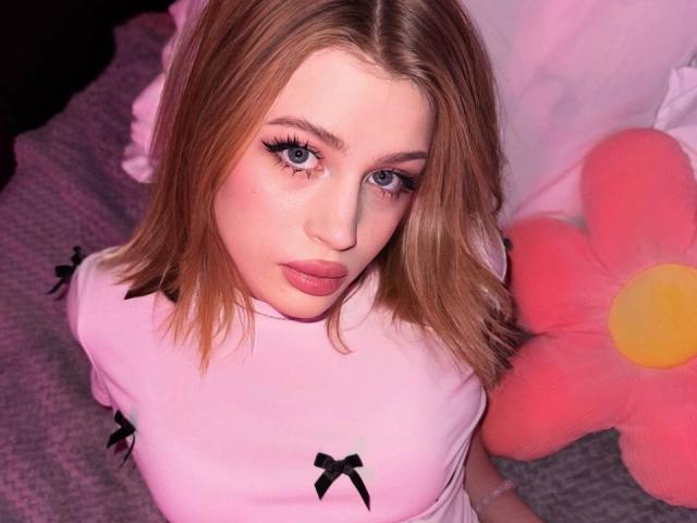 PeiMrozoski, met blond haar, blauwe ogen en roze lippenstift, draagt een roze shirt met zwarte strikken en kijkt met een lichte frons omhoog naar de camera. Haar borsten zijn klein..