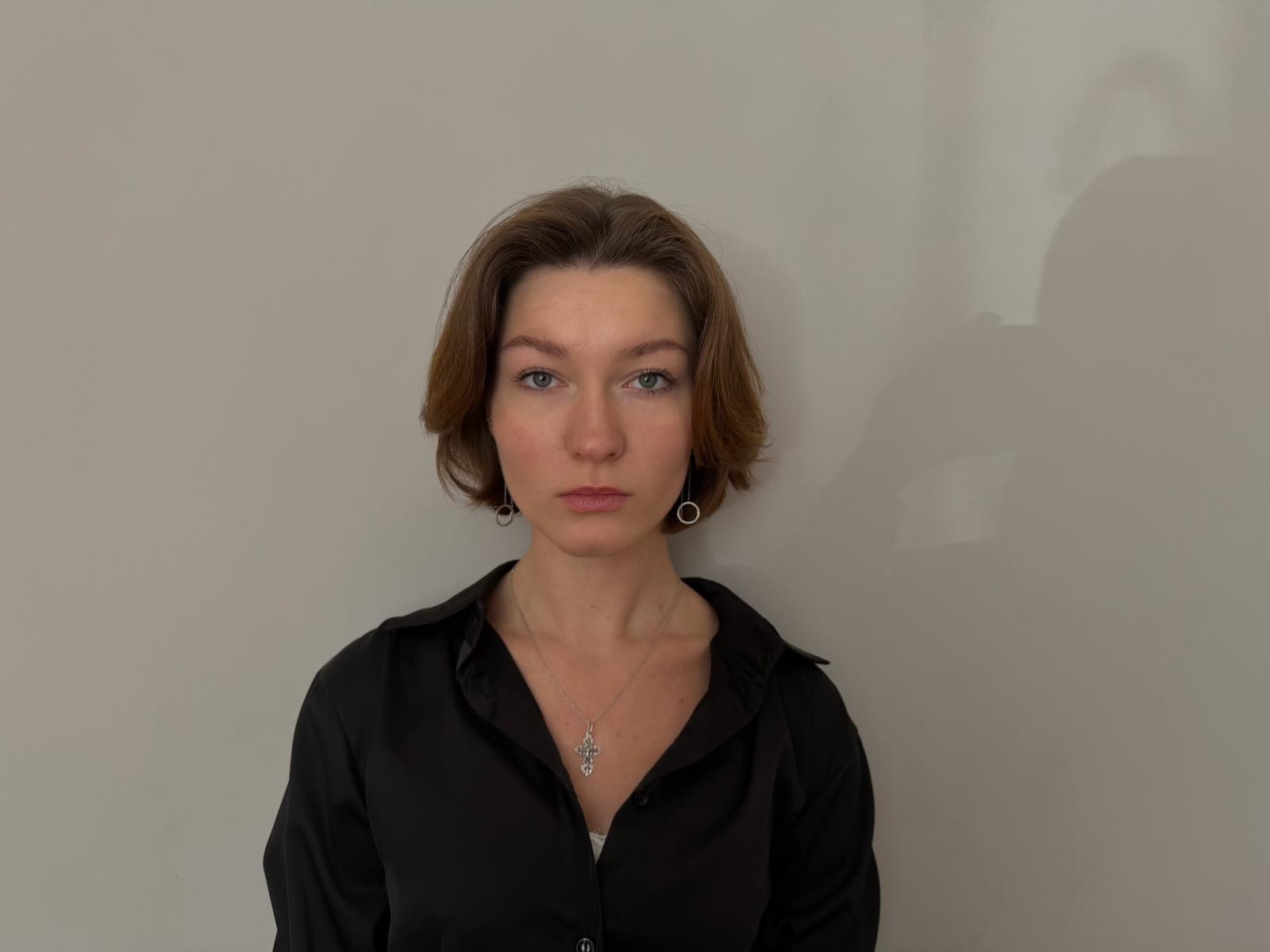 ShantaNewgent, avec des cheveux bruns courts, une chemise noire à boutons, de petites boucles d'oreilles et un collier, regardant droit devant avec une expression neutre, avec une ombre sur la droite..