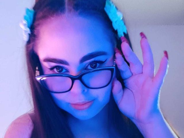 LolaDominatrix, con la piel ligeramente azulada, gafas negras y uñas rojas, sosteniendo su oreja, tiene el cabello largo y castaño, y lleva pinzas con flores, sonriendo con los labios ligeramente entreabiertos..