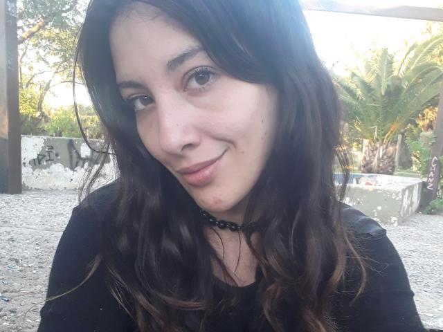 PoppyPlayme, con cabello castaño ondulado, piel clara y una sonrisa sutil, vestida con una blusa negra y un collar de cuentas, mostrando un pequeño grano rojo en su barbilla..