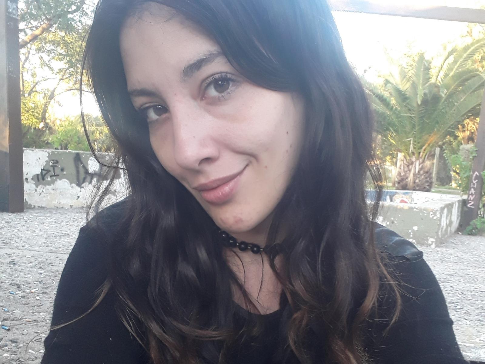 PoppyPlayme with wavy brown hair, light skin, and a subtle smile, wearing a black top and bead necklace, showing a small red pimple on her chin.