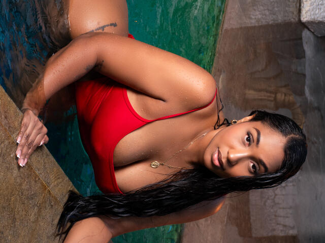 NaiomiLanne with wet black hair, wearing a red one-piece, leaning on a pool edge, showing her tits, small tattoo on her arm, looking at the camera.