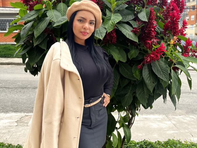 JessicaFiore stands in front of green leaves and red flowers, wearing a beige hat, black shirt, beige coat, and gray high-waisted pants, with a beige belt.