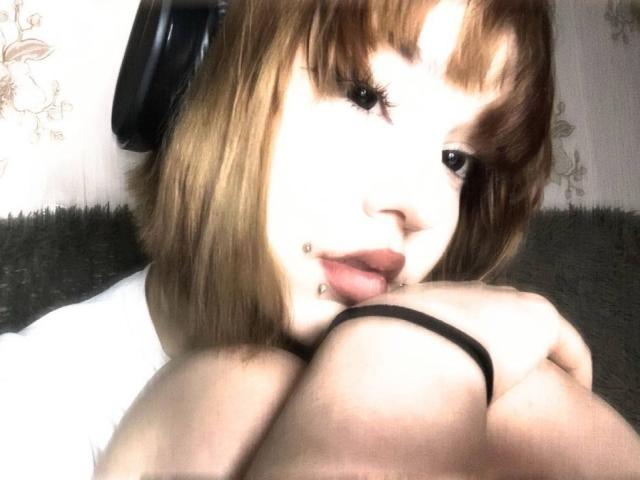 DinahLackey with short brown hair, black headphones, lip piercings, and a black wristband, sitting with knees up, looking at the camera, in a soft, blurry photo.